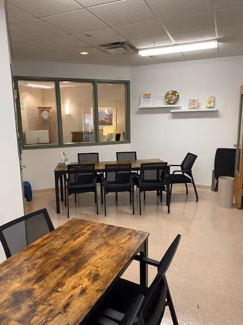 Interior room with two wooden tables and several black chairs arranged around them. There is a window on the left wall showing another room with a grandfather clock and a lamp. On the right wall, a white shelf holds decorative plates and small framed pictures. The floor is light-colored and the ceiling has recessed lighting.