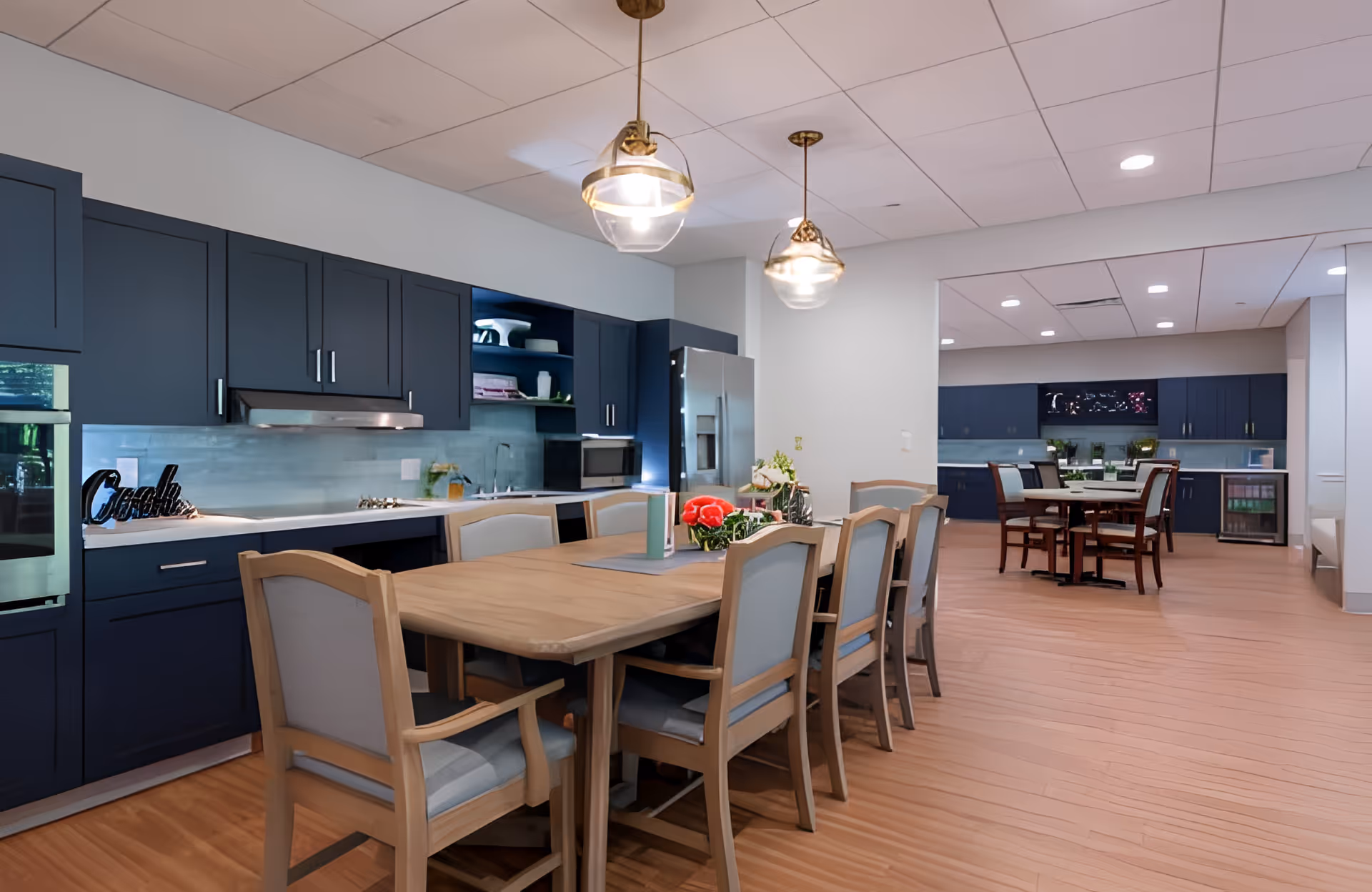 A spacious kitchen and dining area with a long wooden dining table surrounded by cushioned chairs. The kitchen features dark blue cabinets, a stainless steel refrigerator, oven, and microwave. Two pendant lights hang from the ceiling above the dining table. In the background, there is another smaller dining table with chairs and additional cabinetry along the wall.