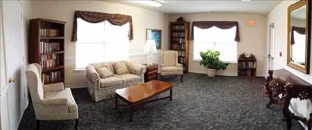 A well-lit living room with two large windows covered by valance curtains. The room contains a beige sofa, two beige armchairs, a wooden coffee table, two bookshelves filled with books, a potted plant, a floor lamp, and a decorative wooden console table with a large mirror above it. The carpet is dark with a subtle pattern.