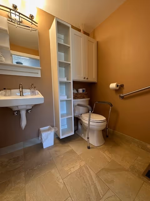 A clean bathroom with beige walls and tiled floor. The bathroom features a white sink with a mirror above it, a white toilet equipped with a metal safety frame, a wall-mounted grab bar, a white trash bin, and a tall white storage cabinet with open shelves and a closed compartment.