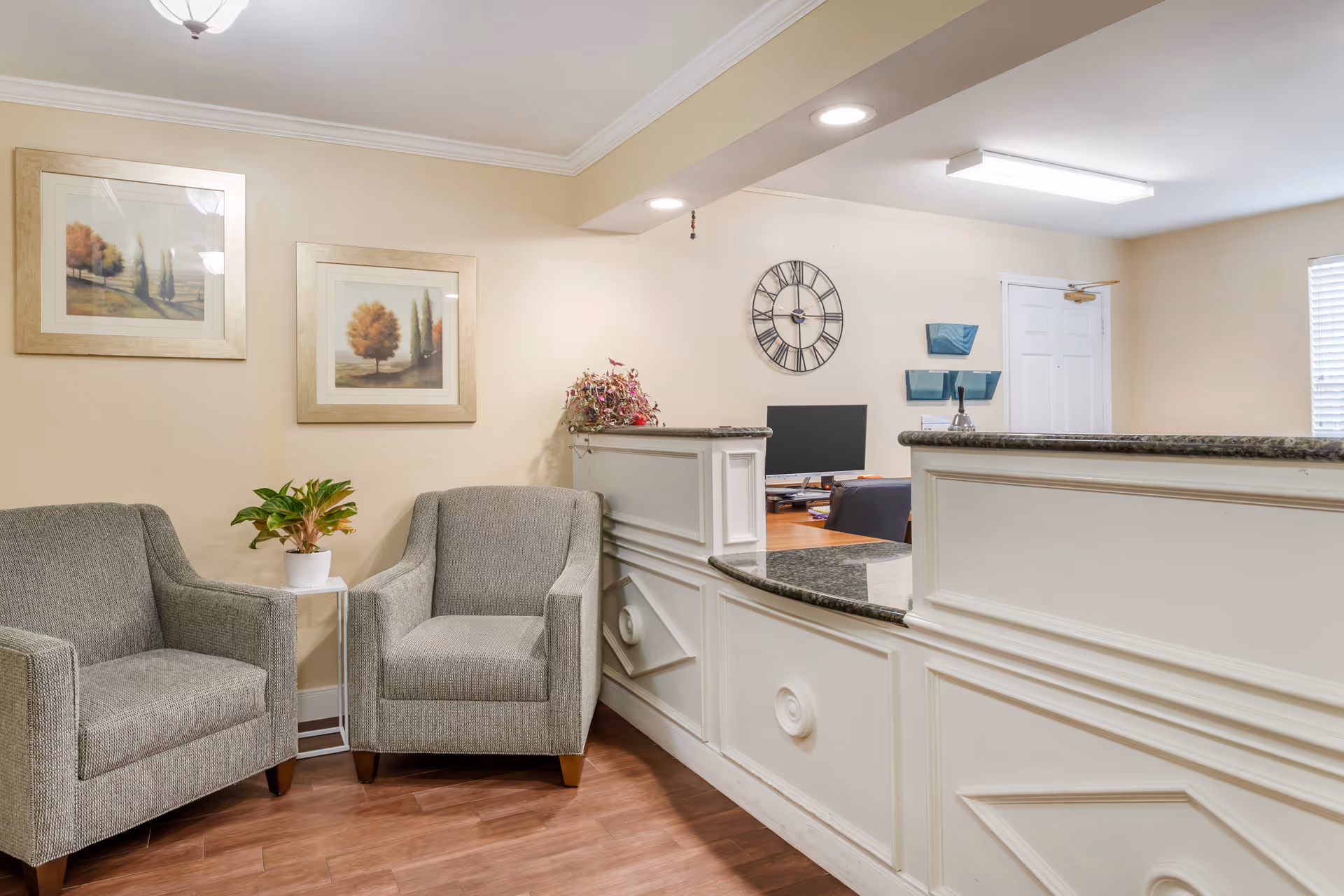 Reception area with a white counter featuring a granite top, two gray upholstered armchairs with a small white side table holding a potted plant between them, two framed landscape paintings on the wall, a large round clock, and a computer on the desk behind the counter.