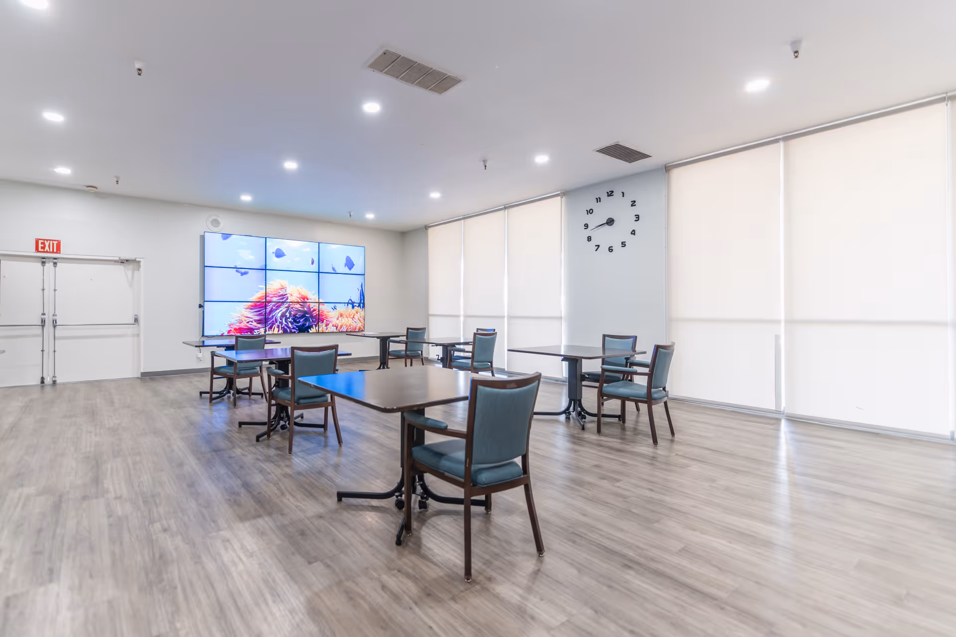 Spacious dining/activity room with several tables and chairs, a large video wall, wall clock, and window shades.