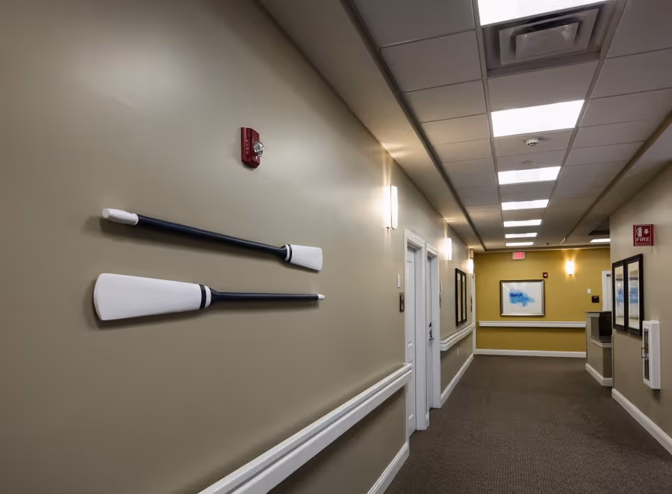 A well-lit hallway in a senior living facility with beige walls and carpeted floor. Two decorative oars are mounted on the left wall. The hallway has several doors, wall-mounted lights, framed artwork, and a fire alarm and extinguisher sign visible.