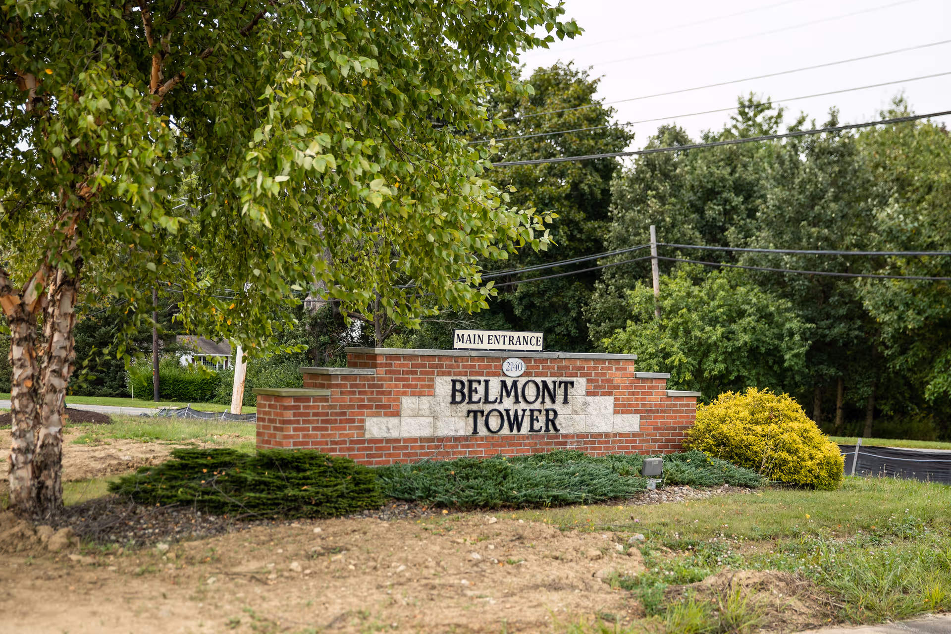Brick entrance sign that reads 'BELMONT TOWER' surrounded by landscaping and trees with a small 'Main Entrance' marker above.
