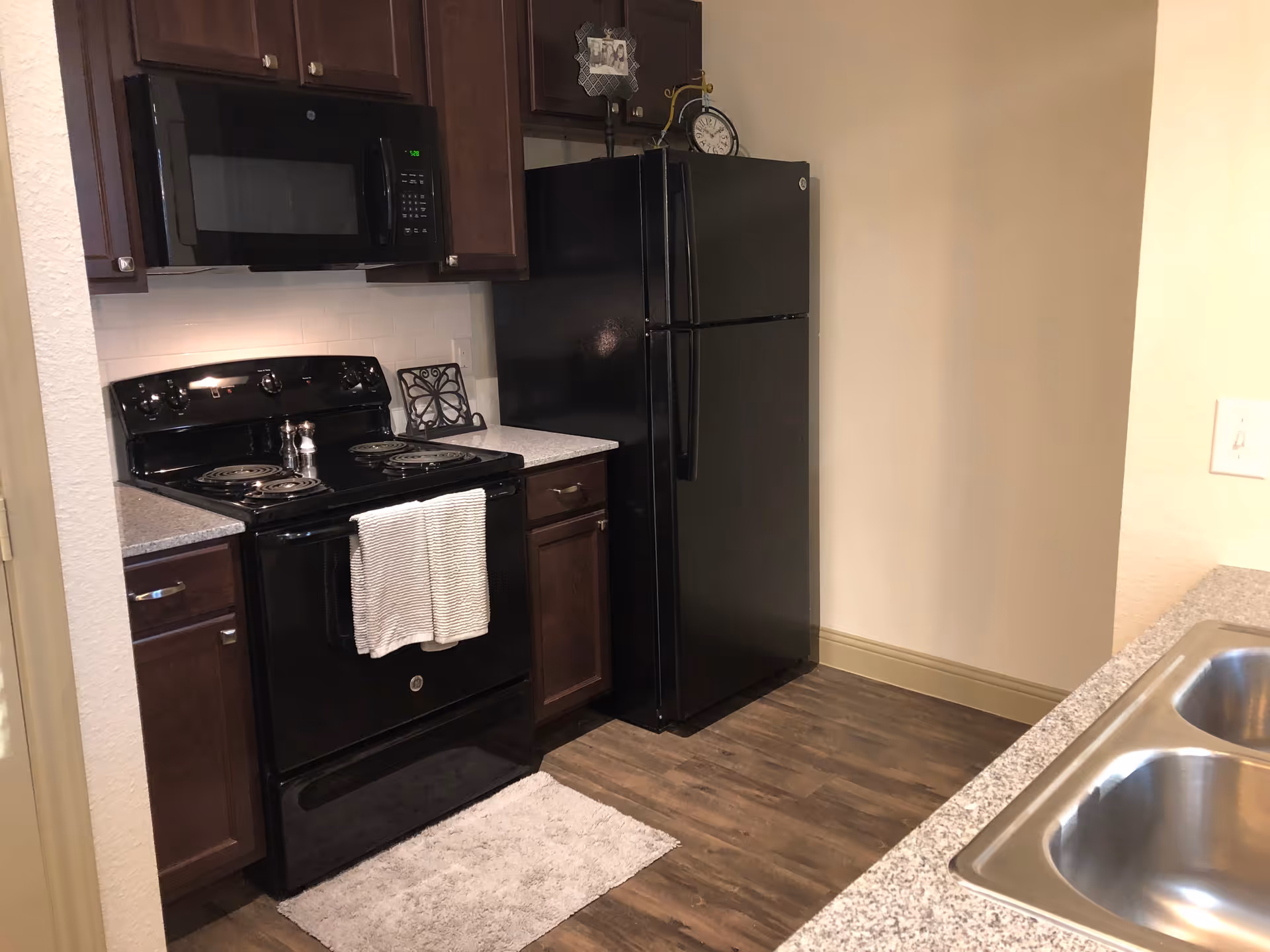 Small kitchen with black stove, microwave, and refrigerator, dark wood cabinets, granite-style counters and a double sink.