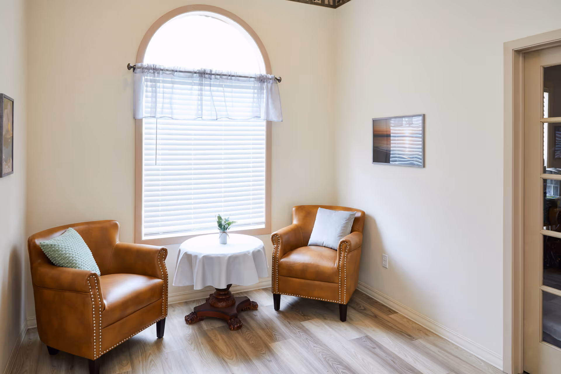 A cozy sitting area with two brown leather armchairs, each with a decorative pillow, positioned around a small round table covered with a white tablecloth. The area is illuminated by natural light coming through a large arched window with white blinds and a sheer white valance. The walls are light-colored, and there is a framed picture hanging on the right wall. The floor is light wood, and a door with glass panels is partially visible on the right side.