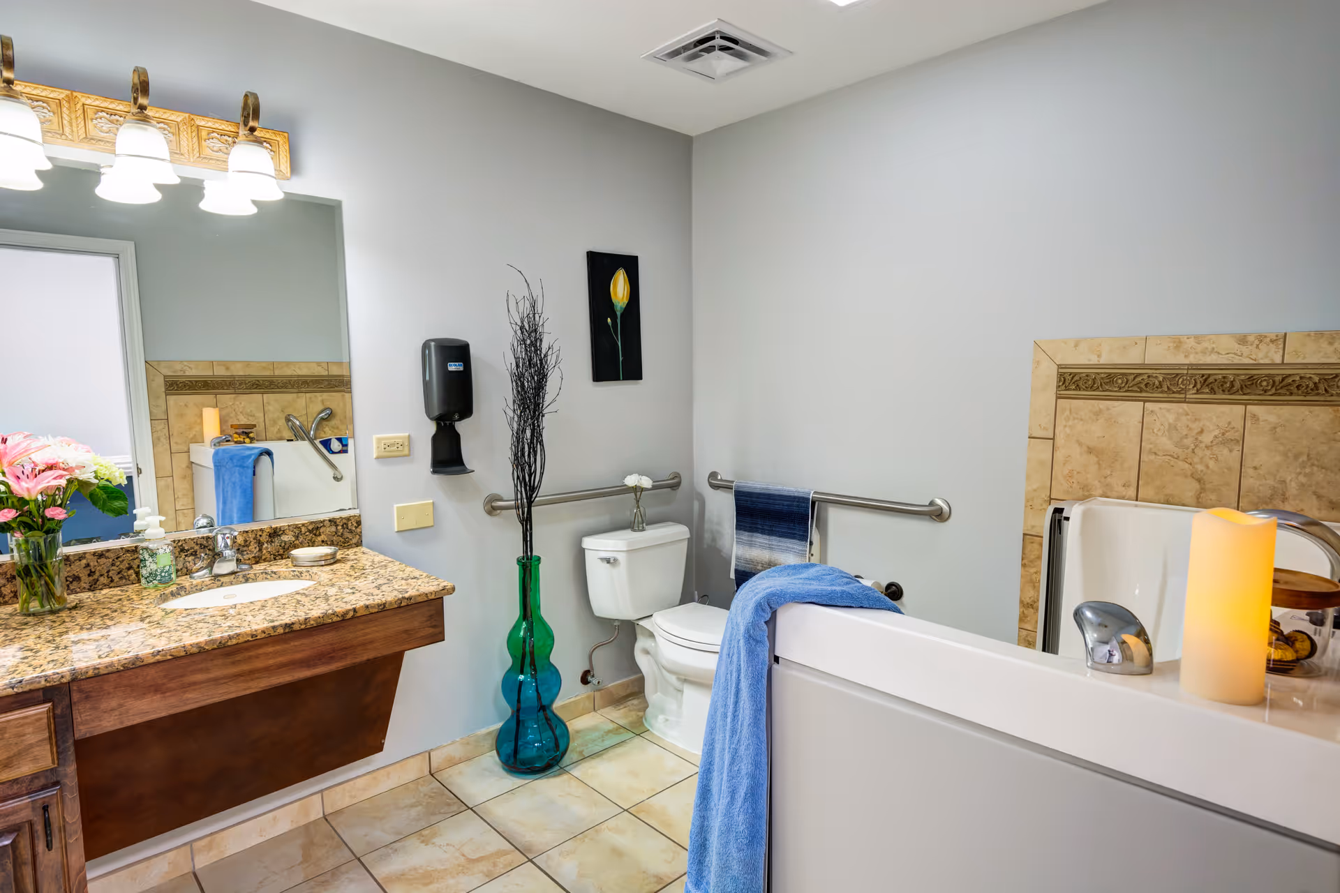 A clean and well-lit bathroom with a granite countertop vanity, a large mirror with lights above, a toilet with grab bars, a blue towel hanging on the grab bar, a decorative tall green vase with branches, a small flower vase on the toilet tank, and a bathtub with a blue towel draped over the side and a lit candle on the edge.