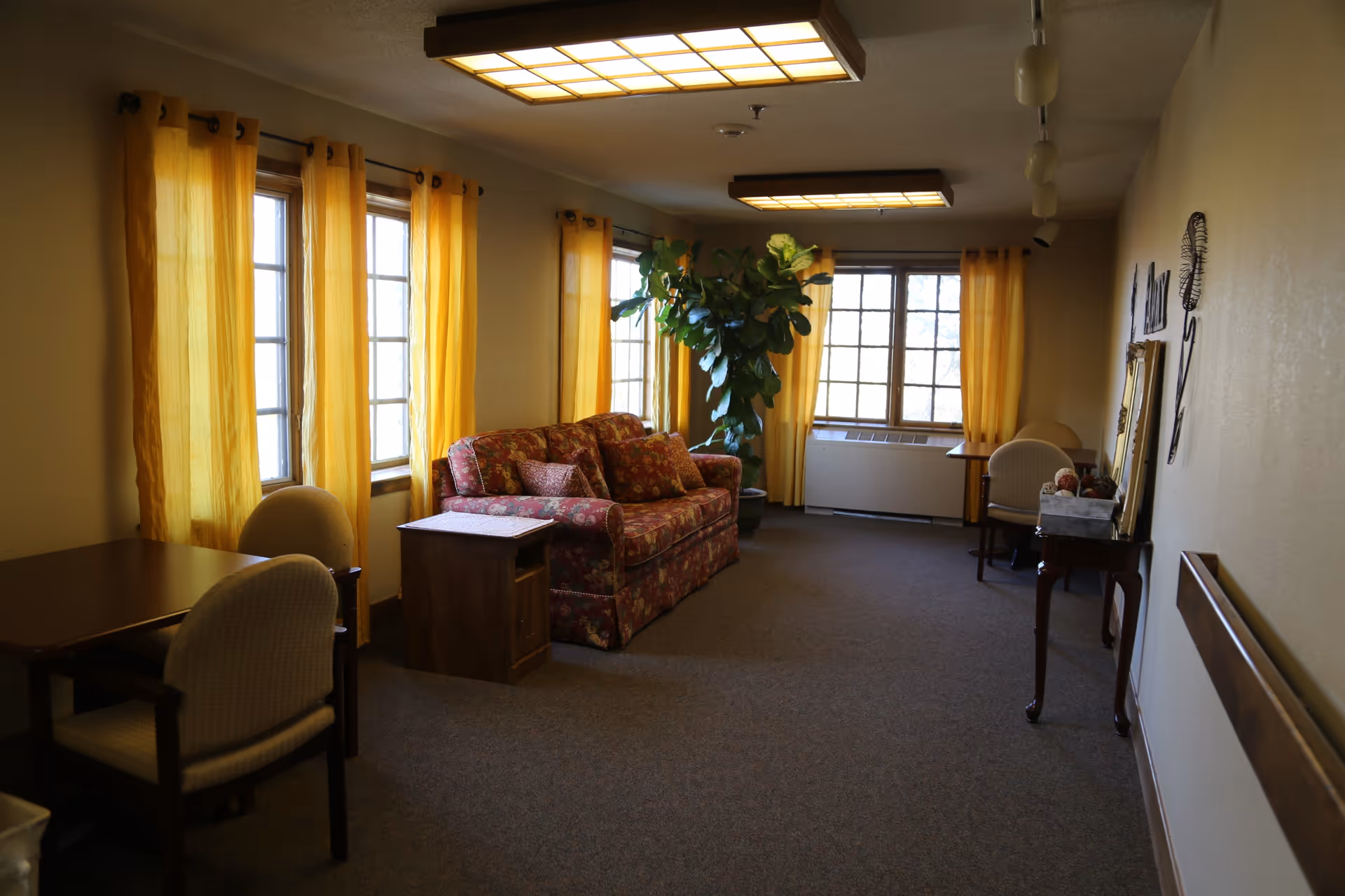 A cozy living room area with yellow curtains on multiple windows, a floral patterned sofa with matching cushions, a large green potted plant, two small tables with chairs, and a decorative console table with a mirror and ornaments on the right wall.
