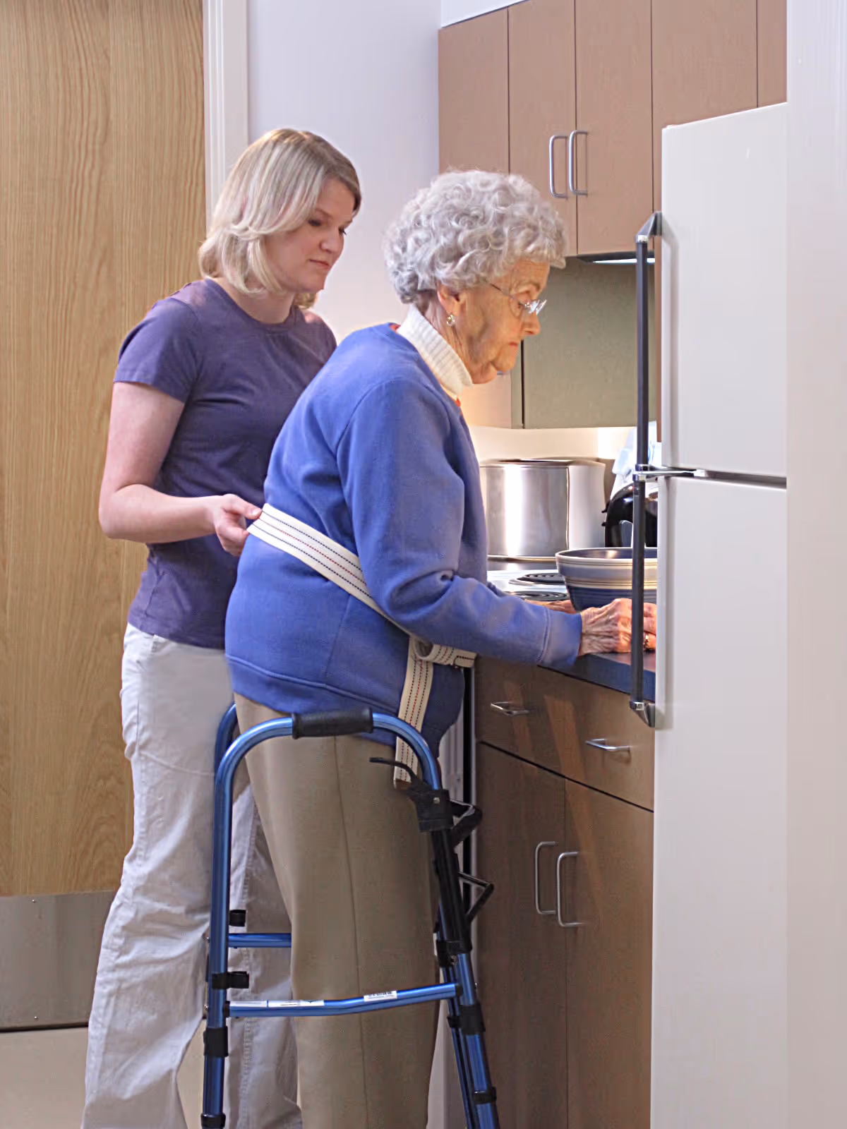 An elderly woman using a walker is assisted by a caregiver in a kitchen area. The elderly woman is wearing a blue sweater and beige pants, and the caregiver is standing behind her, helping with a safety belt around her waist. They are near a countertop with kitchen cabinets and a refrigerator.
