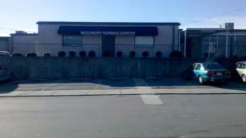 Exterior view of Woodbury Nursing Center with a fence and parked cars.