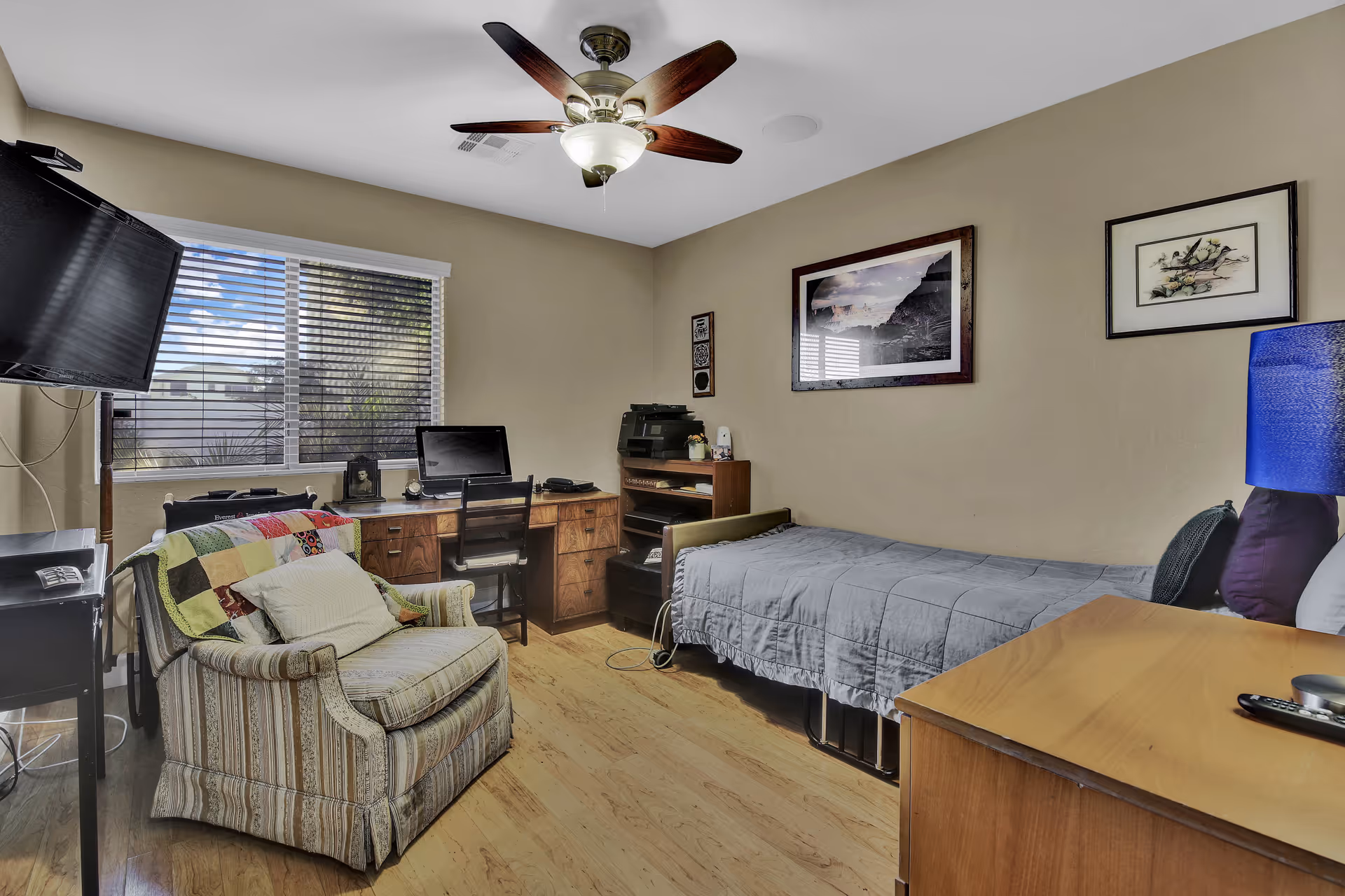 A cozy bedroom with a single bed covered in a gray quilt, a wooden desk with a computer and printer, a striped armchair with a colorful patchwork blanket, a wall-mounted TV, and framed artwork on beige walls. The room has a ceiling fan with lights and a window with blinds letting in natural light.