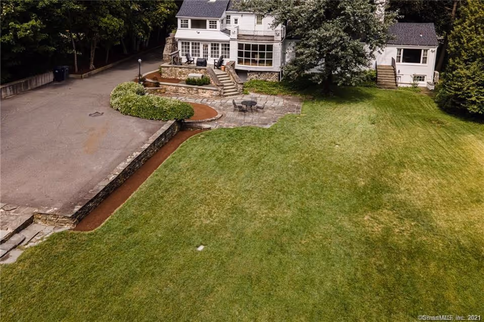 Aerial view of a residential property with a large green lawn, stone patio with outdoor furniture, a driveway, and a white multi-story house surrounded by trees.