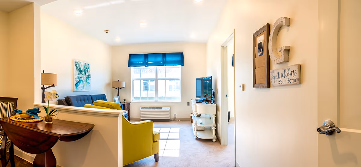 Interior view of a senior living facility's common area at Demaree Crossing featuring a small living room with a blue sofa, a green armchair, a wooden side table with a TV, and a window with a blue valance letting in natural light. A small dining table with chairs and place settings is partially visible on the left. The walls are decorated with artwork and a welcome sign.