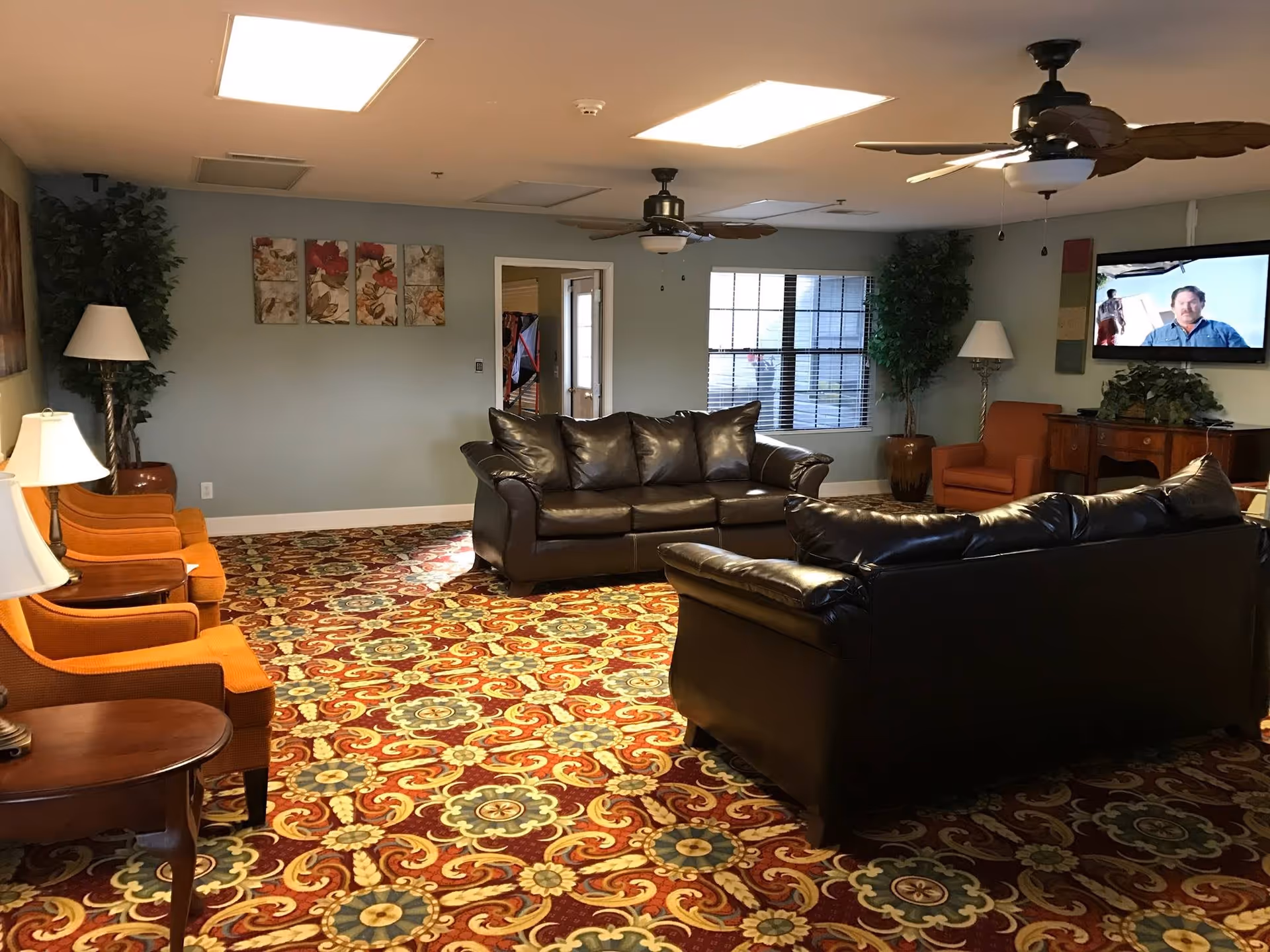 A cozy living room in an assisted living facility with two black leather sofas, three orange armchairs, two floor lamps, a patterned carpet with red, yellow, and blue floral designs, ceiling fans, a wall-mounted TV showing a man, and decorative plants and artwork on the walls.