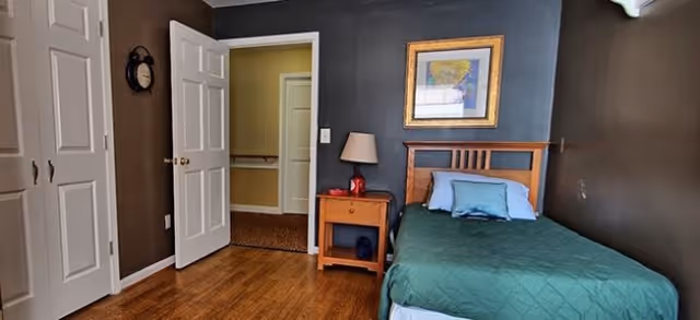 A small bedroom with a single bed covered in a green quilt and blue pillows. Next to the bed is a wooden nightstand with a lamp and a red decorative item. The walls are painted dark brown, and there is a framed picture hanging above the bed. The room has wooden flooring and a white door that is open, showing a hallway with beige walls and carpeted floor.