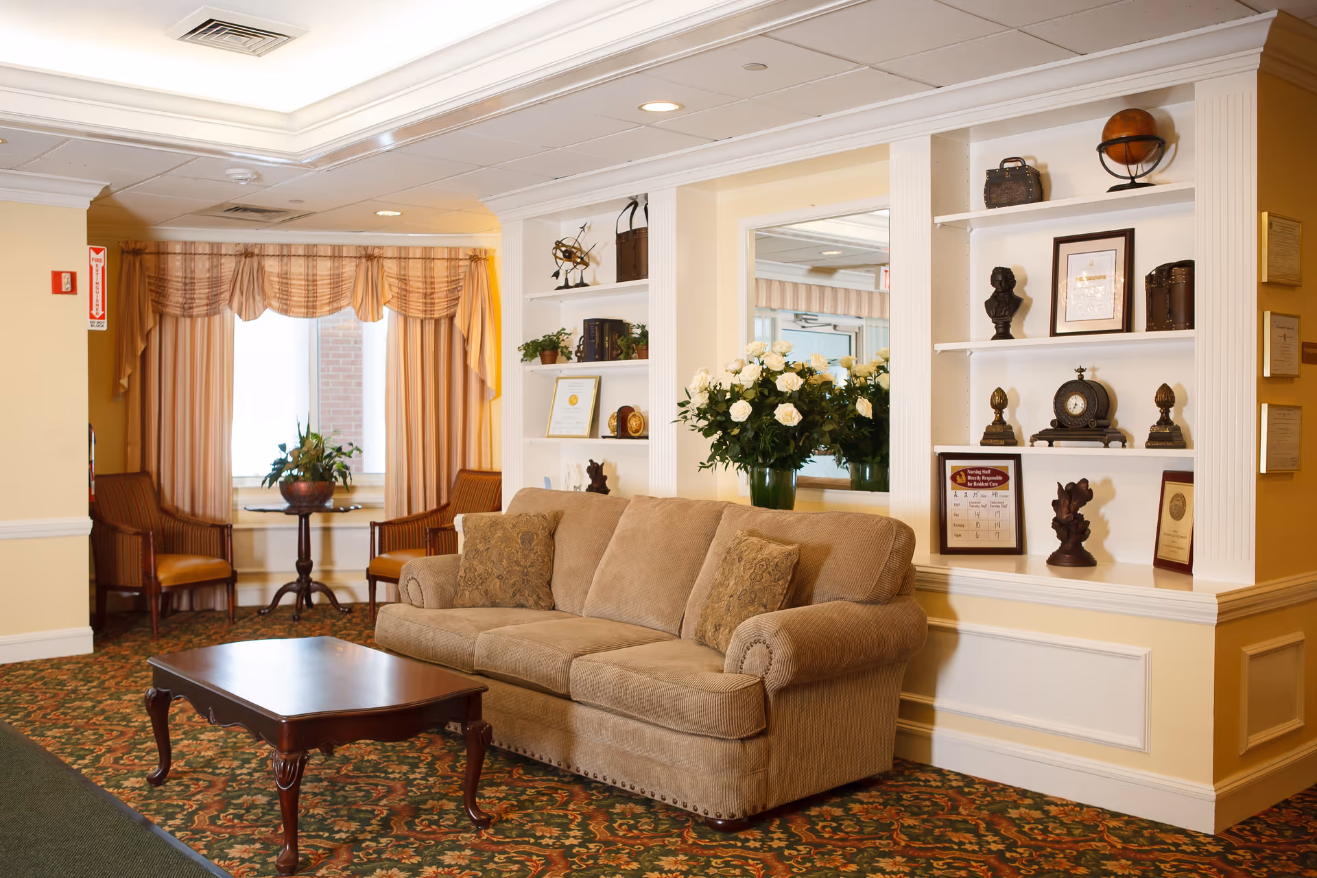 A cozy living room area with a beige sofa adorned with two patterned cushions, a dark wooden coffee table in front, and two wooden chairs with orange cushions near a window with beige and orange drapes. Built-in white shelves on the right display various decorative items including a clock, framed certificates, a bust, and a globe. A vase with white roses sits on a ledge behind the sofa. The room has warm yellow walls and a patterned carpet.