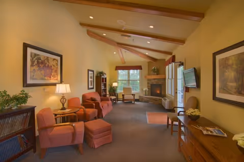 Community living room with upholstered armchairs and ottomans, a fireplace under a vaulted beamed ceiling, bookshelves, and a wall-mounted TV.