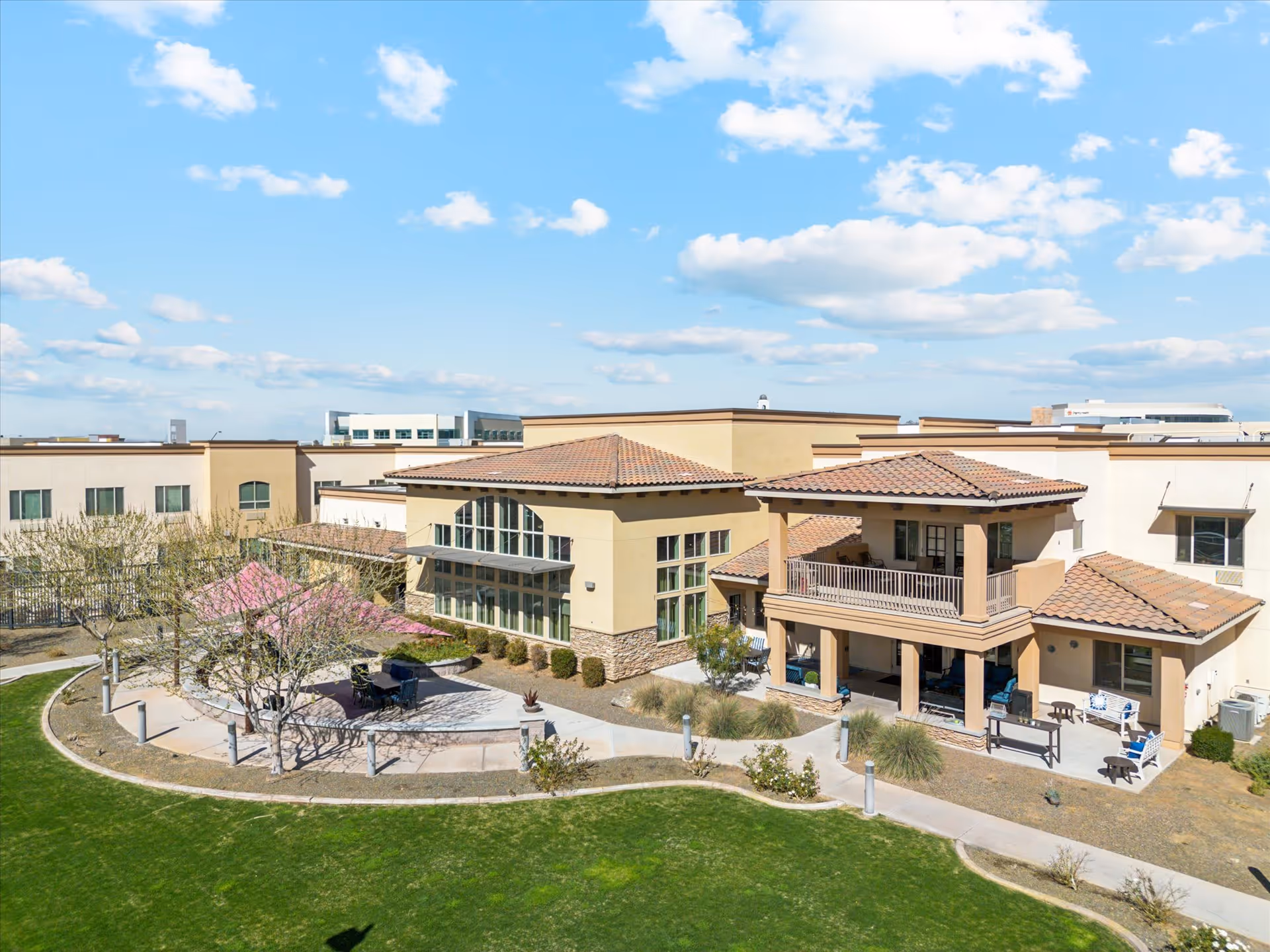 Exterior view of a senior living facility building with beige walls and red-tiled roofs under a blue sky with scattered clouds. The building has large windows and a covered patio area with outdoor seating. There is a curved walkway and landscaped greenery including grass, bushes, and trees in the foreground.