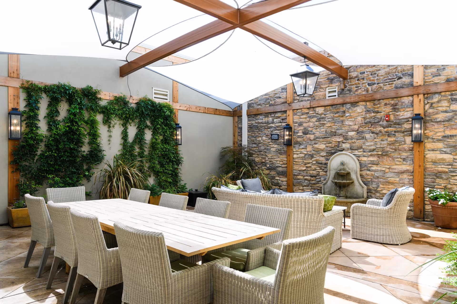 Covered outdoor courtyard with a long wooden dining table, wicker chairs and lounge seating against a stone wall with a fountain and climbing plants.