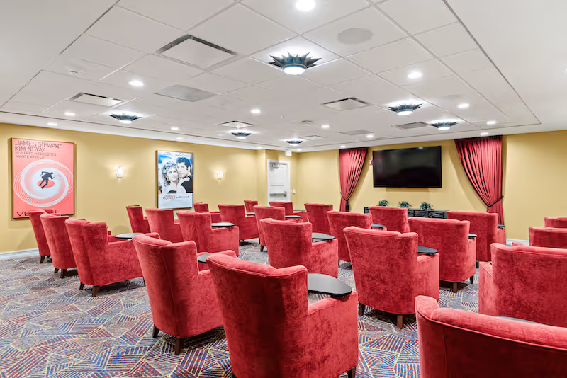 A cozy theater room with multiple rows of red upholstered armchairs, each equipped with a small black side table. The room has a patterned carpet, yellow walls adorned with movie posters, a large flat-screen TV mounted on the wall, and red curtains framing the TV. The ceiling has recessed lighting and decorative light fixtures.