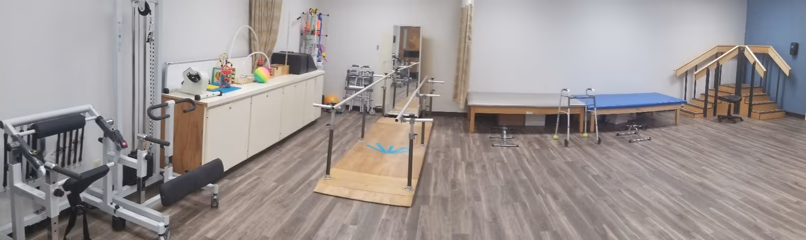 A spacious physical therapy or rehabilitation room with various exercise and therapy equipment including parallel bars, a small staircase with handrails, therapy tables, and exercise machines. The room has wood-patterned flooring and light-colored walls.