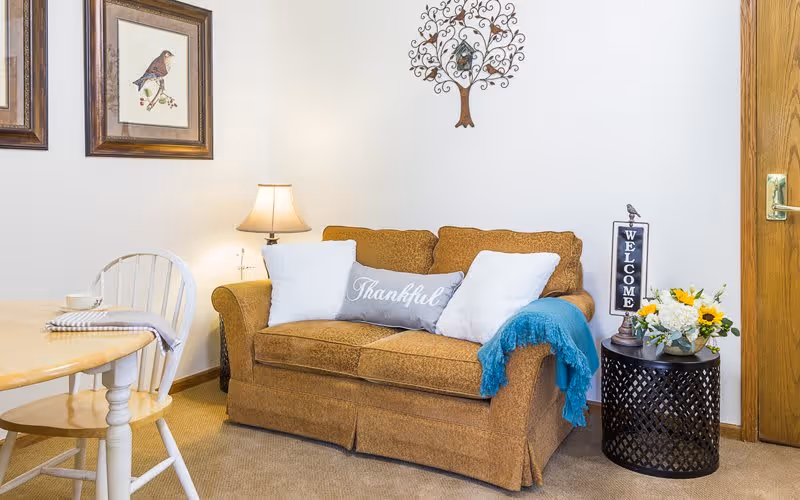 A cozy living room area with a brown loveseat adorned with three pillows, one gray with the word 'Thankful' and two white. A blue throw blanket is draped over one arm of the loveseat. To the right is a round black side table with a 'WELCOME' sign, a small bird figurine, and a floral arrangement with sunflowers. On the left, there is a wooden dining table with a white chair and a table lamp on a small stand behind the loveseat. The walls are decorated with two framed bird pictures and a metal tree wall art.
