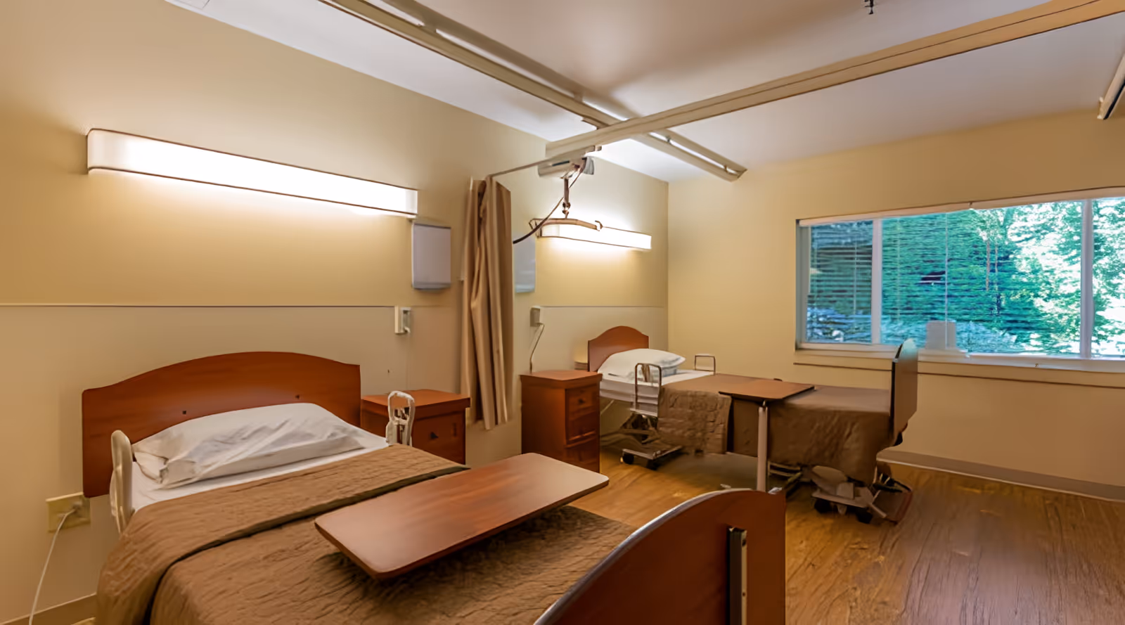 A rehabilitation health center room with two hospital beds, each equipped with a brown blanket and a wooden overbed table. The room has beige walls, wooden flooring, and a large window showing greenery outside. There are wall-mounted lights above each bed and a privacy curtain between the beds.