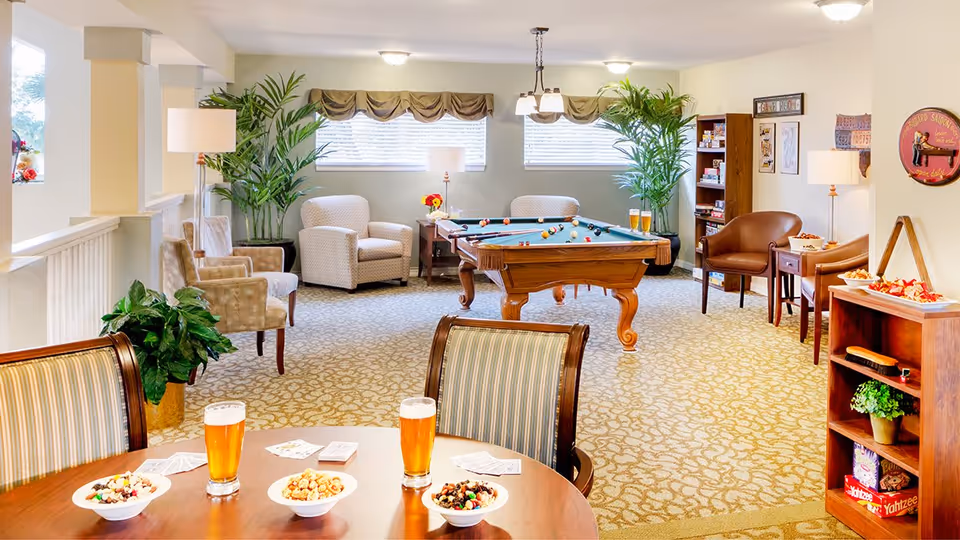 A bright and inviting common area with a pool table in the center, surrounded by comfortable armchairs and plants. There is a round table in the foreground with snacks and glasses of beer. The room has large windows with blinds and valances, carpeted floor, and shelves with board games and decor.