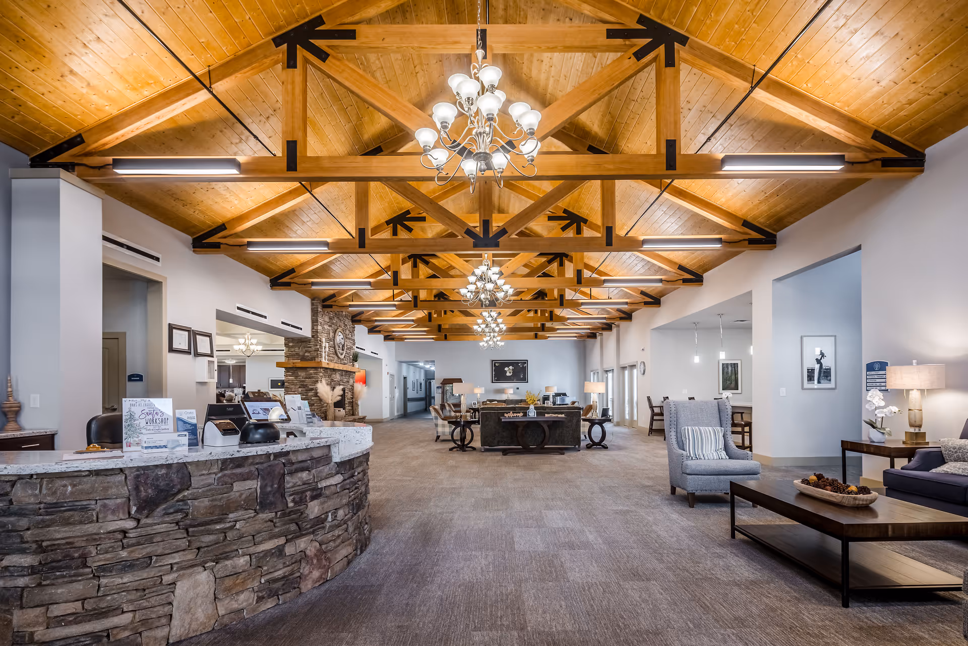 Spacious senior living lobby with a stone reception desk, exposed wooden beam ceiling, chandeliers, and multiple seating areas.