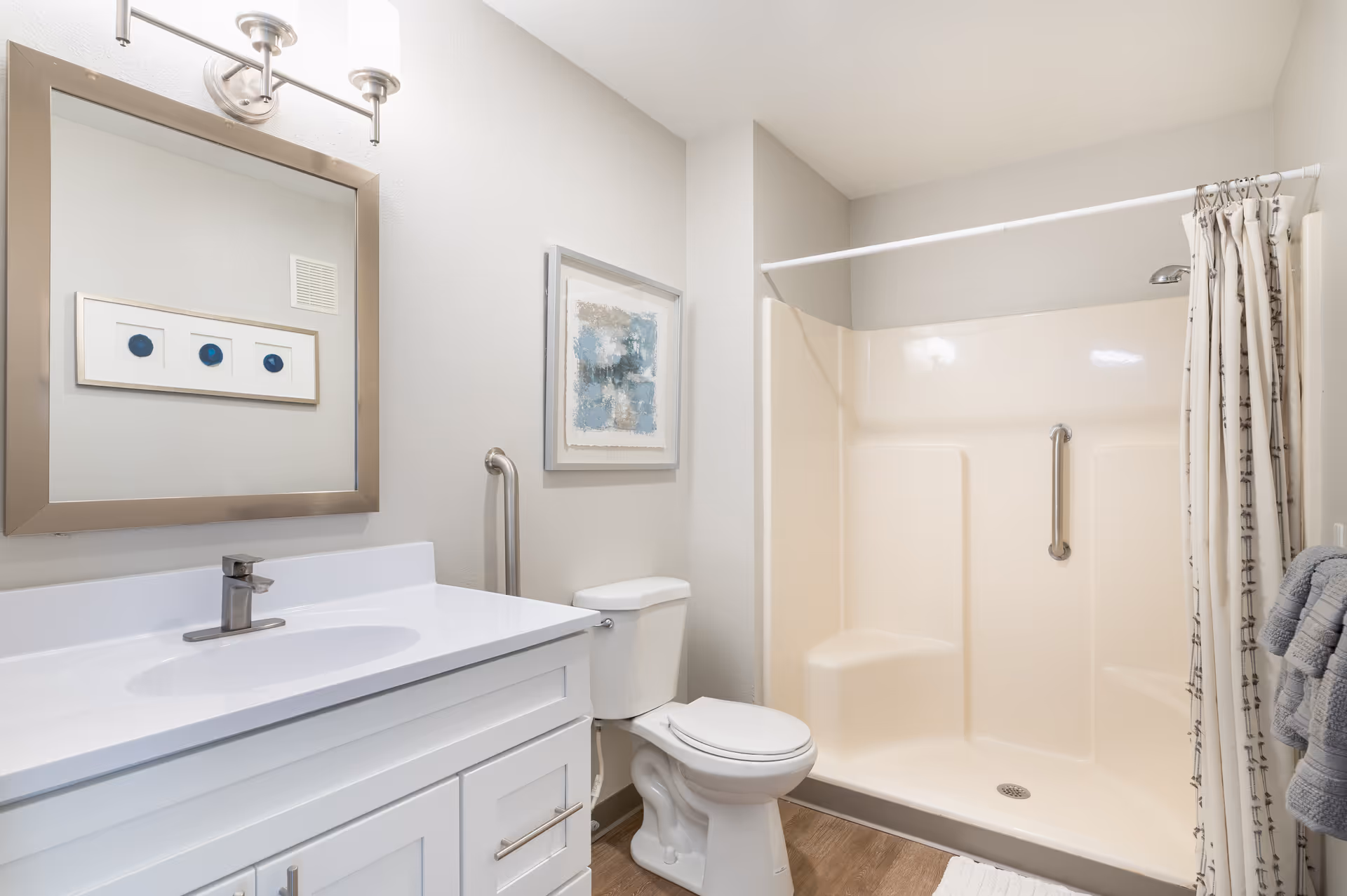 A clean and modern bathroom featuring a white vanity with a sink and faucet, a large mirror with a silver frame, a toilet, and a walk-in shower with a built-in seat and grab bars. The walls are light-colored, and there is a framed abstract artwork hanging above the toilet. Towels are neatly hung on a rack beside the shower.