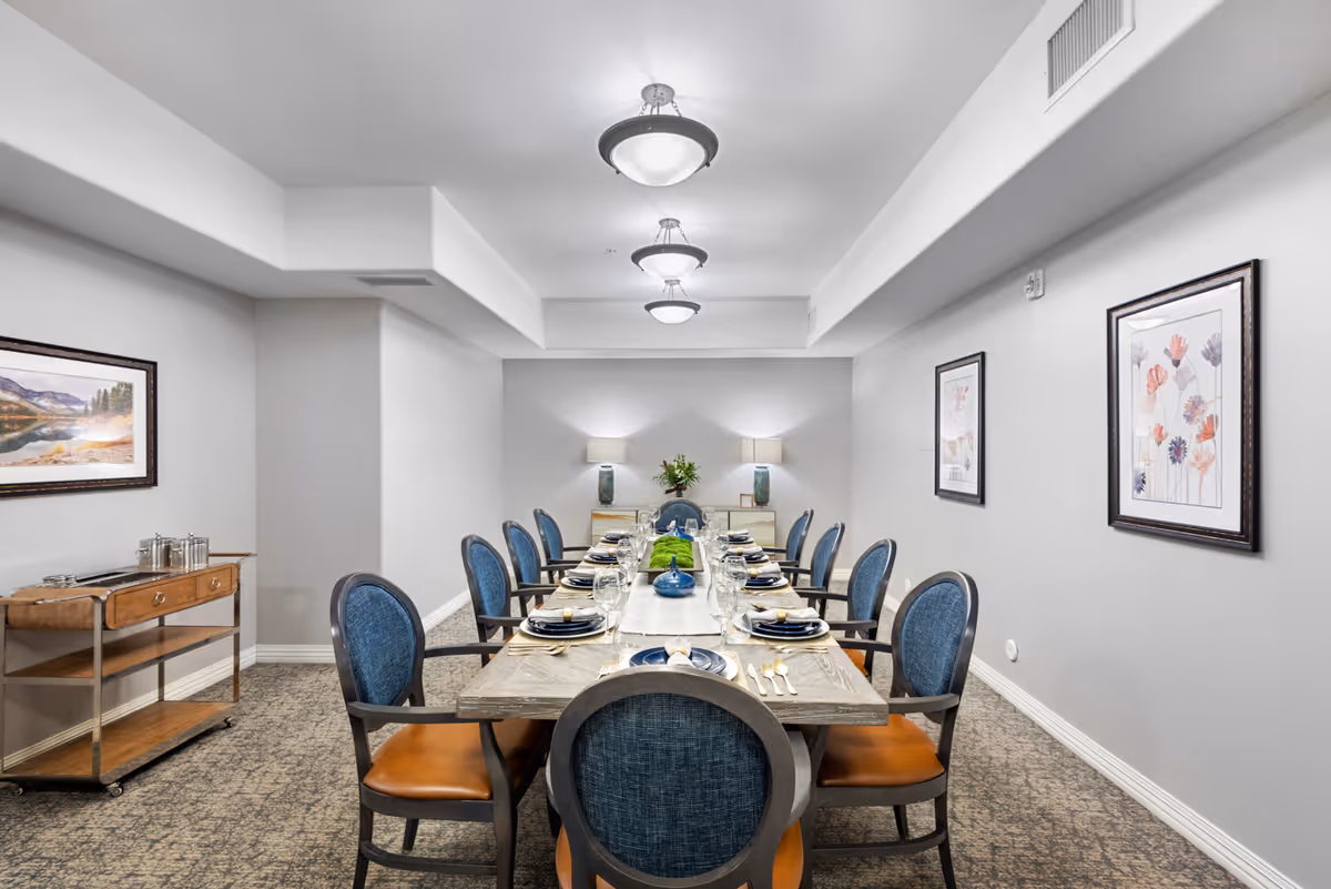 A long dining table set for a meal with blue cushioned chairs around it in a well-lit room. The table is decorated with plates, glasses, cutlery, and a centerpiece of greenery. The room has light gray walls adorned with framed floral artwork and a landscape painting. There is a wooden serving cart on the left side and two table lamps on a sideboard at the far end of the room.
