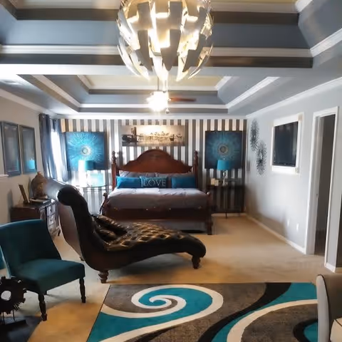 A spacious bedroom with a large wooden bed featuring a 'LOVE' pillow, two blue lamps on bedside tables, a chaise lounge chair, a teal armchair, and a modern chandelier hanging from a coffered ceiling. The room has striped wallpaper behind the bed and a colorful abstract rug on the floor.