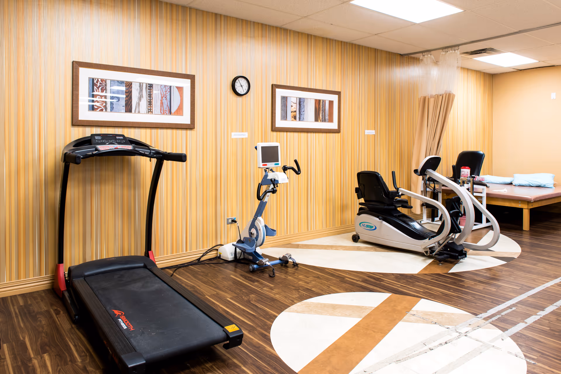 A fitness room with a treadmill, a stationary exercise bike, and a recumbent exercise bike. The room has wood-patterned flooring with a circular design, striped wallpaper, two framed pictures on the wall, a clock, and a treatment table with pillows and a curtain in the corner.
