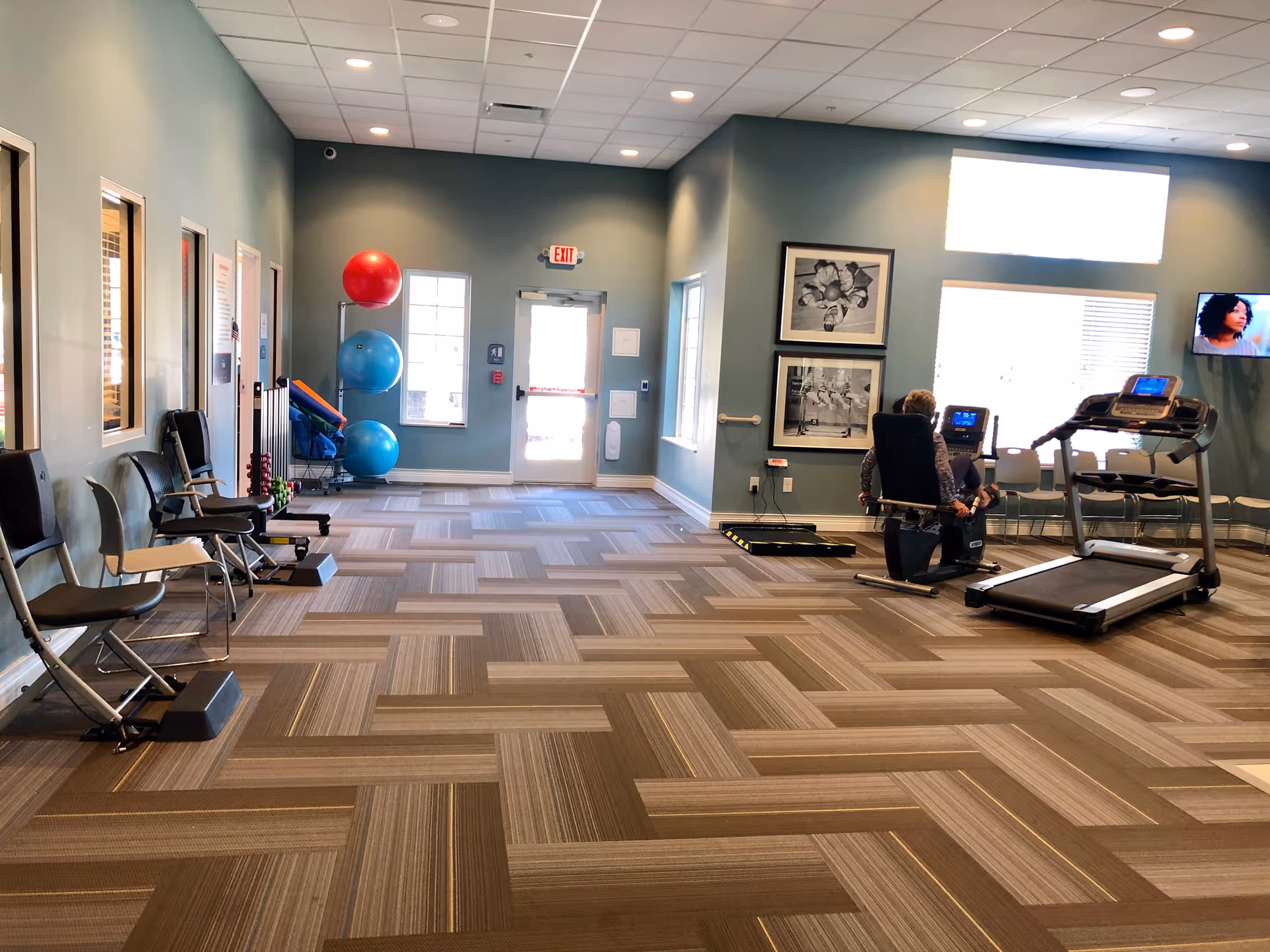 Bright senior fitness room with treadmills, exercise machines, chairs and stacked exercise balls, and a person using a recumbent bike.