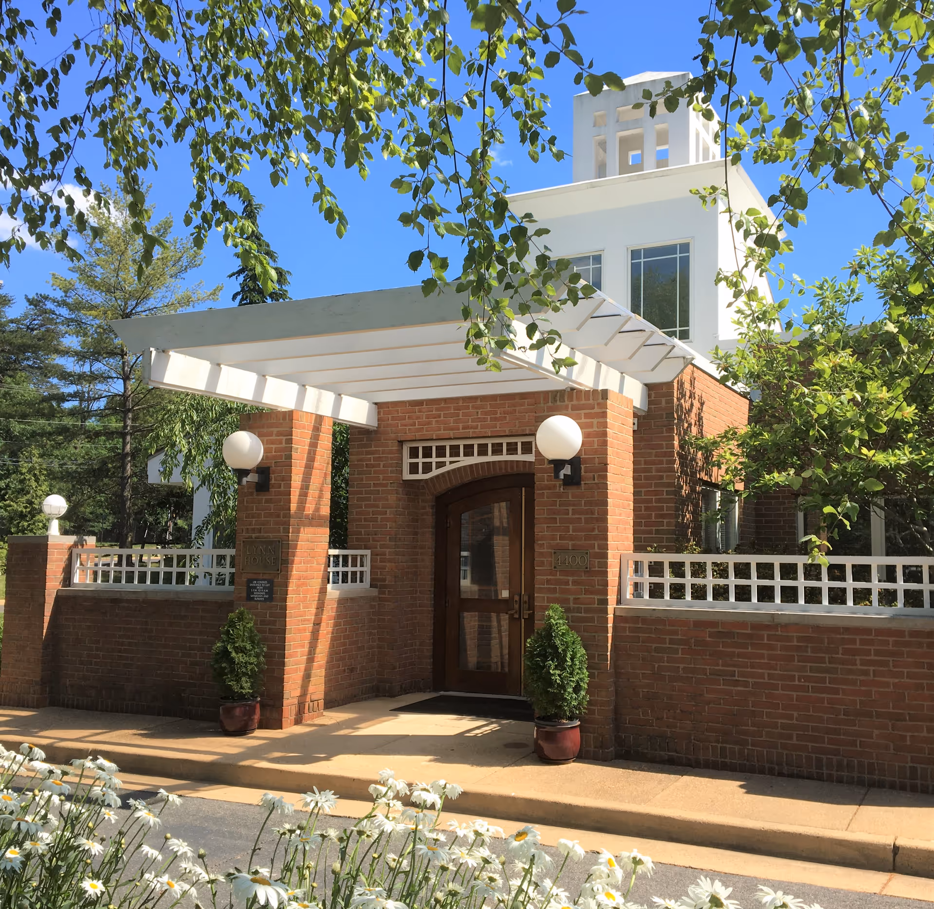 Exterior view of Lynn House-Potomac Valley, showing a brick building entrance with a white pergola-style awning, two round white light fixtures on either side of the door, potted plants, and a clear blue sky with green tree branches framing the scene.