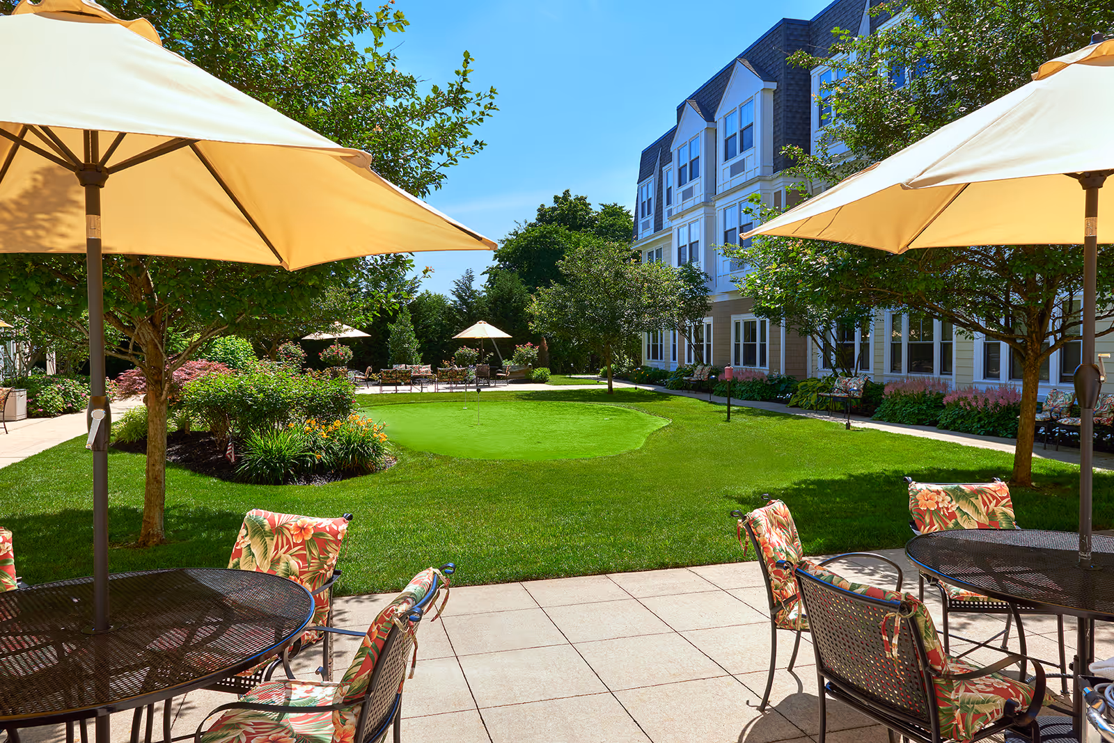Outdoor patio area with tables and chairs under large beige umbrellas, overlooking a well-maintained green lawn with a putting green and surrounded by trees and shrubs. In the background, there is a multi-story residential building with many windows.