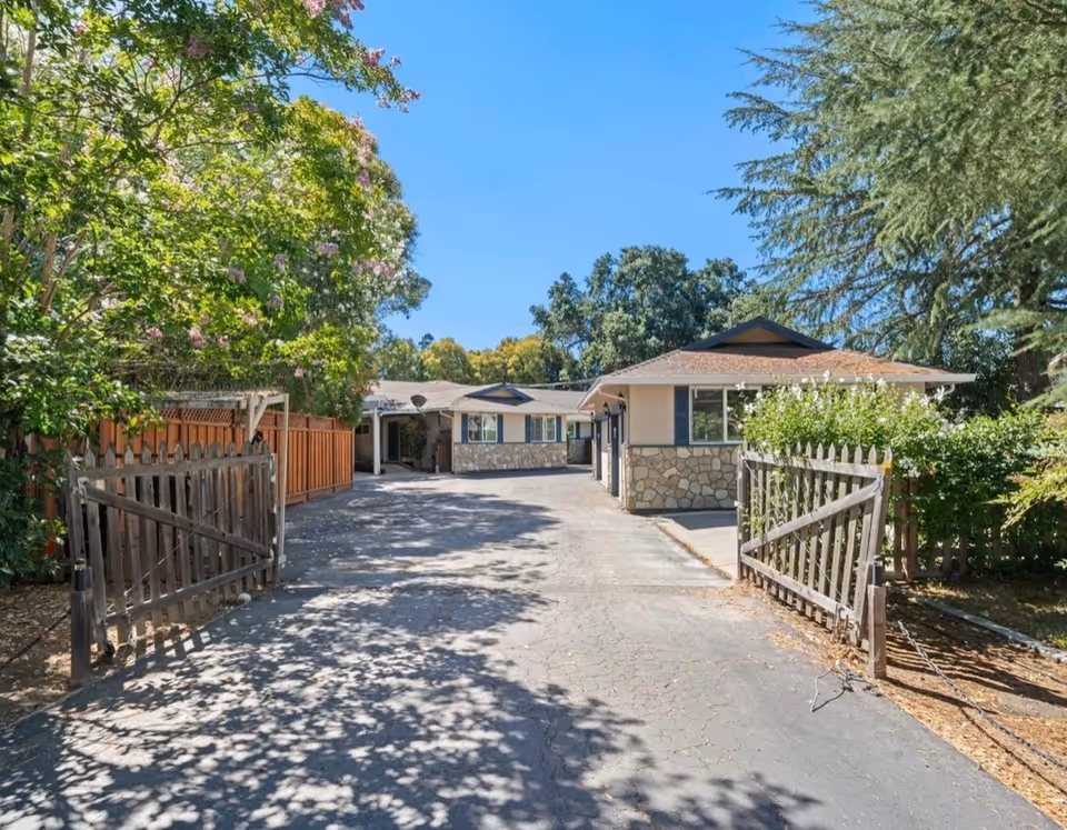 Driveway entrance with open wooden gates leading to a single-story stone-faced building surrounded by trees under a blue sky.