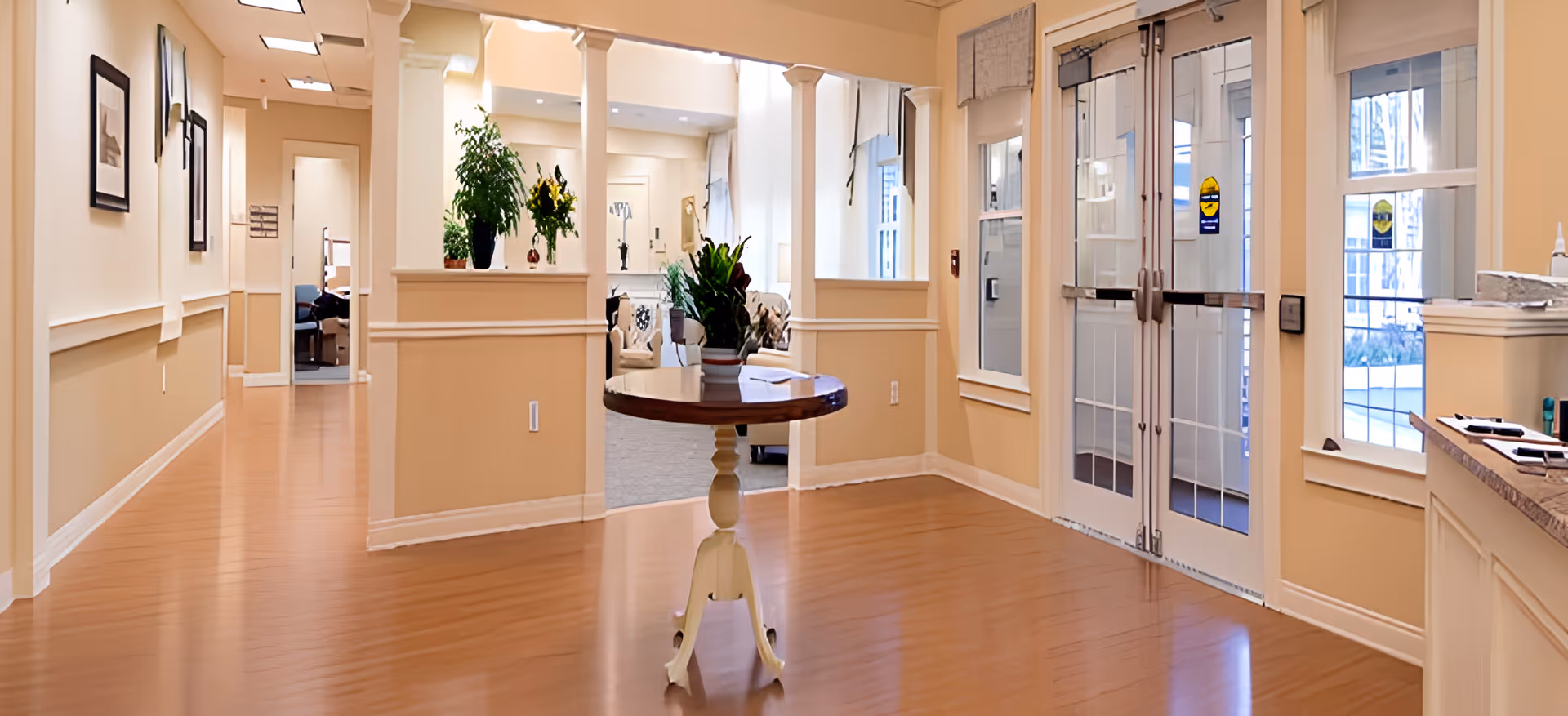Interior view of a senior living facility hallway with light beige walls and wooden flooring. A round wooden table with a potted plant is centered in the foreground. The hallway leads to a bright common area with seating and more plants. Double glass doors with white frames and windows are on the right side, allowing natural light to enter.