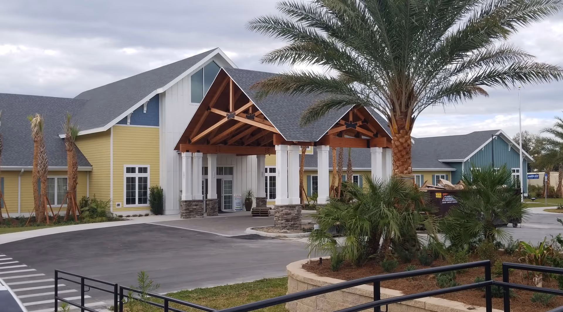 Exterior view of a senior living facility building with a large covered entrance supported by white columns and wooden beams. The building has yellow and blue siding with white trim and a gray roof. There are palm trees and landscaped greenery around the entrance and parking area under a cloudy sky.