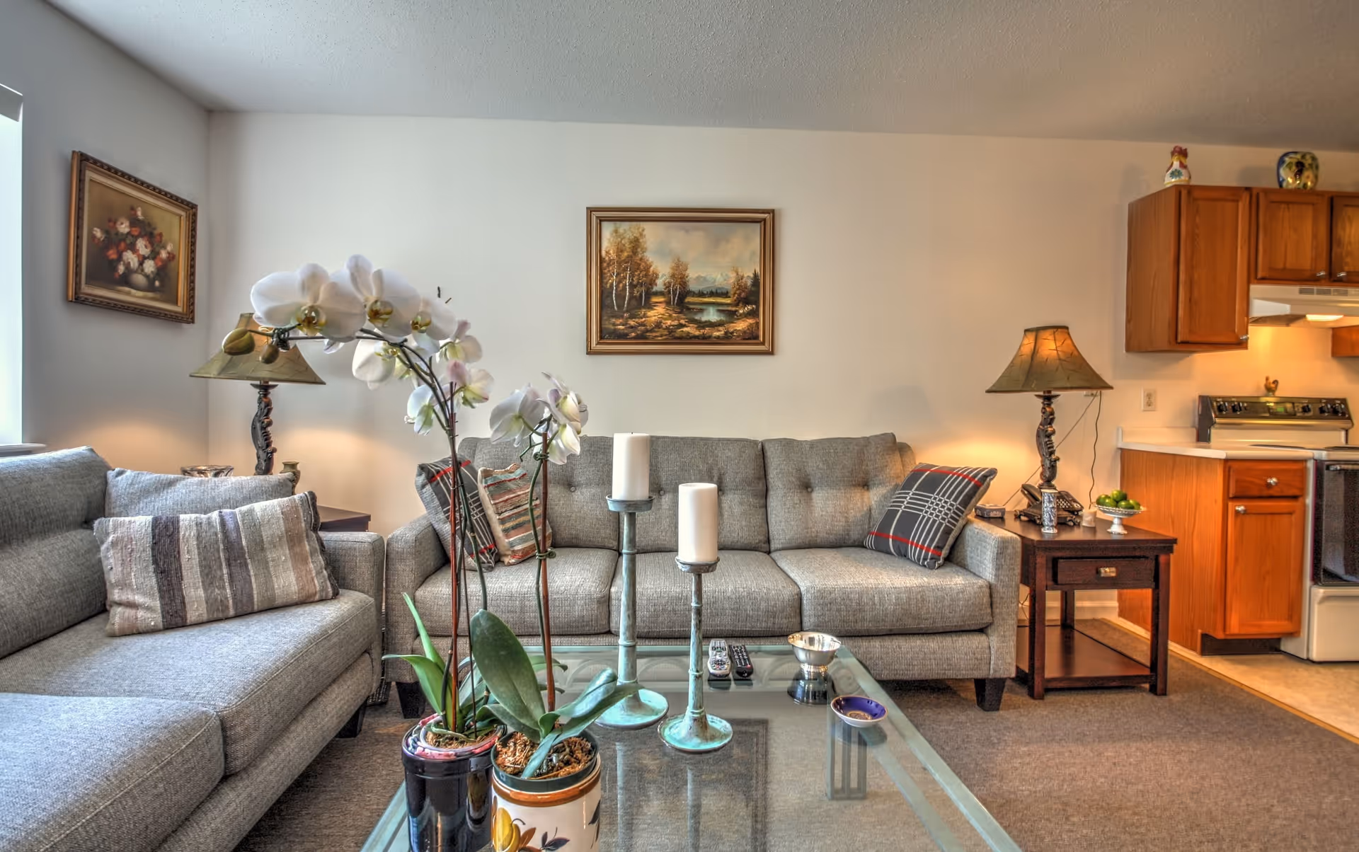 A cozy living room area with two gray sofas adorned with decorative pillows, a glass coffee table holding two potted white orchids and two candle holders with white candles. The room features a side table with a lamp and a telephone, a painting of a landscape on the wall, and a partial view of a kitchen with wooden cabinets and a stove.