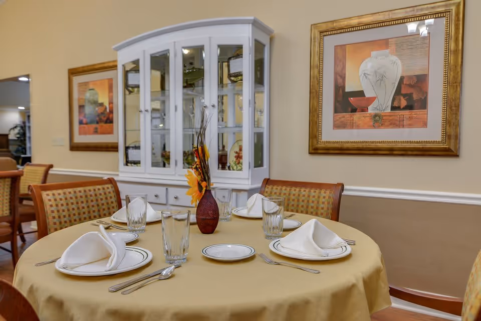 A round dining table set for four with white plates, folded napkins, glasses, and silverware on a yellow tablecloth. A small vase with orange flowers is in the center. Behind the table is a white china cabinet with glass doors displaying decorative items, and framed artwork hangs on the beige walls.
