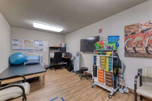 A therapy or rehabilitation room with a black treatment table, a blue exercise ball, and a blue pillow on the left. There is a desk with a black office chair, computer, and printer against the back wall. On the right side, there is a wall-mounted TV above a cart holding colorful therapy weights and exercise equipment. Two chairs are positioned along the walls, and a painting of bicycles hangs on the right wall. The room has wood flooring and white walls with fluorescent lighting.