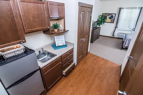 View of a small kitchen area with wooden cabinets, a stainless steel sink, a mini refrigerator, and a basket with bread and eggs on the counter. The kitchen opens into a living space with a dresser, a TV, a plant, a framed picture on the wall, and a bed near a window with blinds and an air conditioning unit.