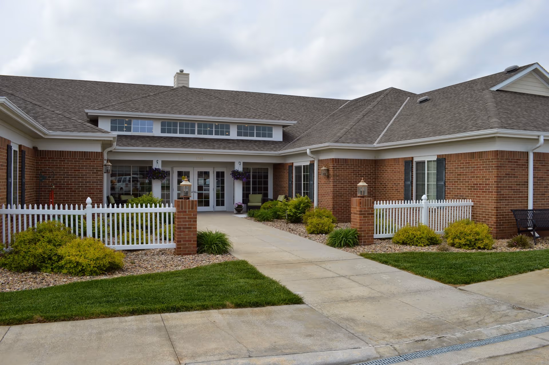 Front exterior view of a single-story brick building with a gray shingled roof, white trim, and large windows. The entrance features double glass doors with sidelights and transom windows above. There are white picket fences on either side of the walkway leading to the entrance, with green shrubs and landscaping around the building. The sky is cloudy.