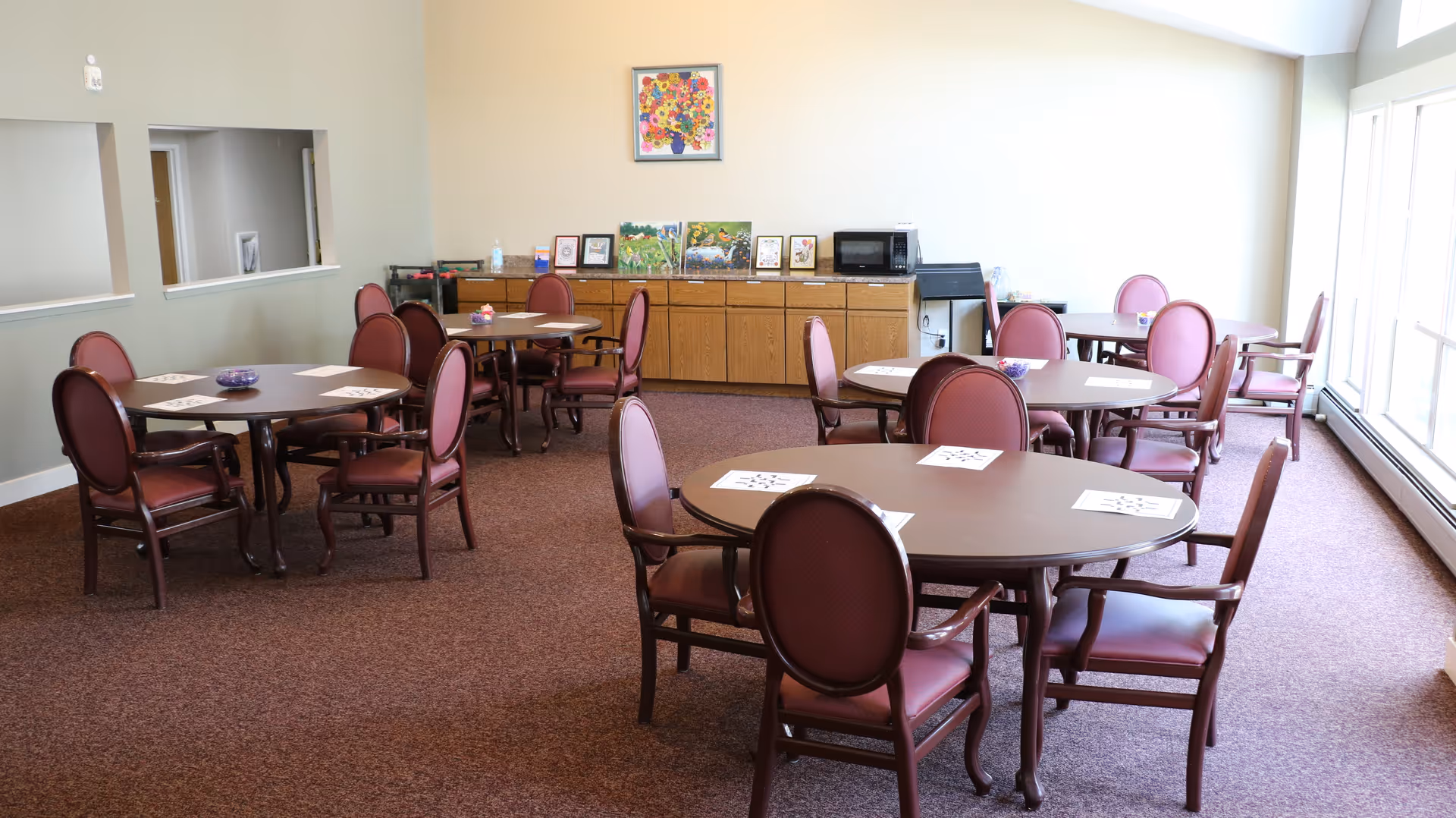 Bright dining room with round tables and burgundy chairs in a senior living facility.