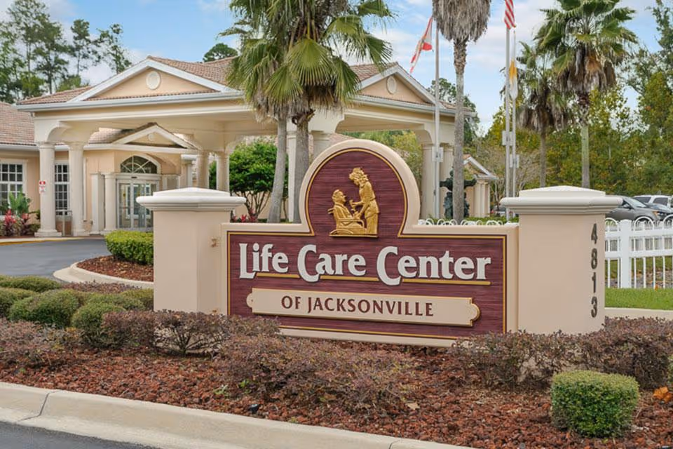 Entrance sign for Life Care Center of Jacksonville with a beige and maroon color scheme, featuring a gold emblem of a caregiver assisting an elderly person. The sign is surrounded by landscaping with bushes and mulch, and the facility building with columns and a covered driveway is visible in the background.