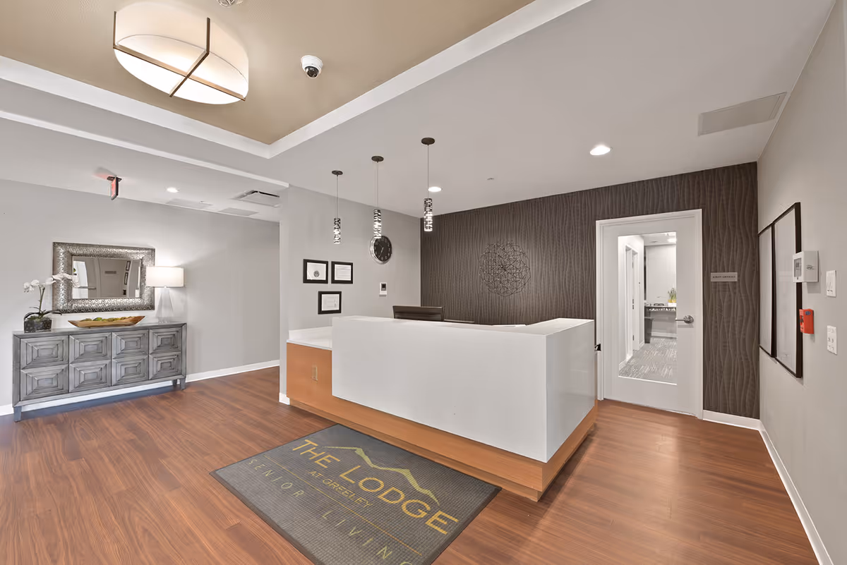 Reception area of The Lodge at Greeley senior living facility with a modern white and wood front desk, a decorative wall with a clock and artwork, a door labeled Staff Offices, a sideboard with a mirror and lamp, and wood flooring.