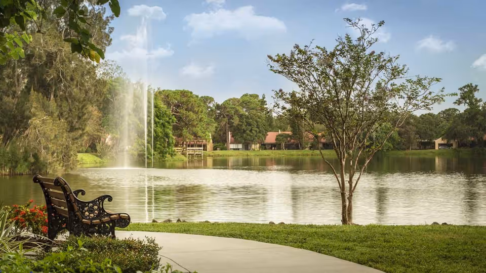 A peaceful outdoor scene featuring a curved pathway next to a lake with a water fountain. There is a decorative bench on the left side near some red flowers and greenery. Across the lake, trees and houses with red roofs are visible under a partly cloudy sky.