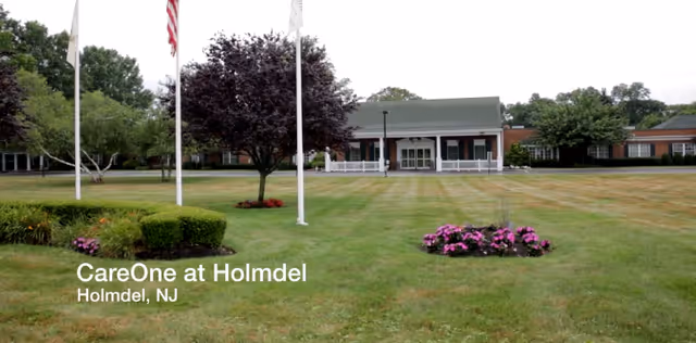 Front exterior view of CareOne at Holmdel facility showing a large green lawn with flower beds, three flagpoles, and a single-story brick building with a covered entrance and white railings in the background.
