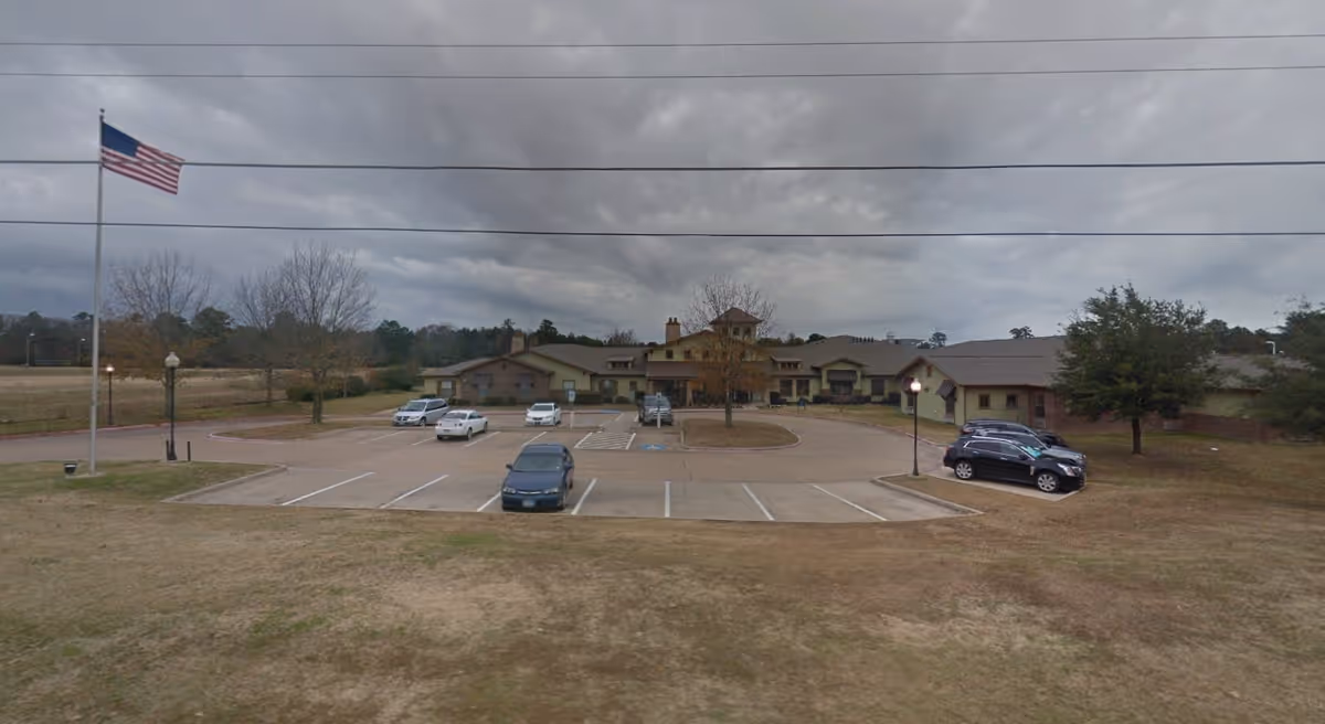 Exterior view of Hawkins Creek Assisted Living and Memory Care facility showing a large building with multiple sections, a parking lot with several cars, an American flag on a flagpole, and a cloudy sky overhead.